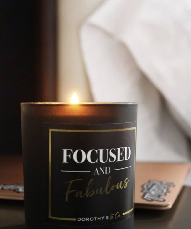 A lit black candle with white and gold text that says 