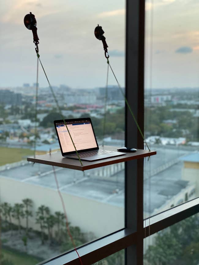 A wooden desk hung up by ropes and suction to a window with a laptop on it 