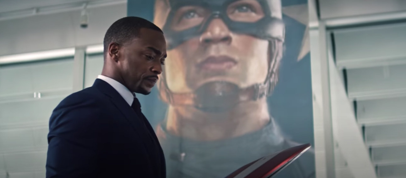 Mackie looks at Captain America&#x27;s shield in front of a Captain America banner in The Falcon and the Winter Soldier