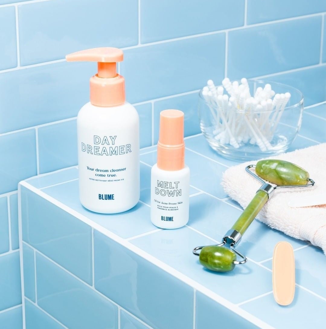 A bottle of cleanser, acne oil, and a jade roller on a tiled bathroom vanity