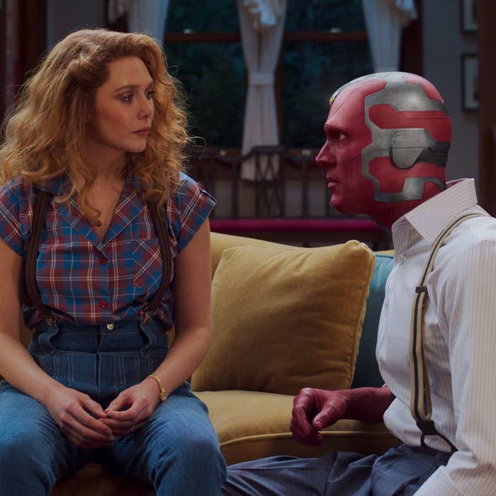 Wanda and Vision sitting on a couch in old time clothing in a sitcom inspired atmosphere 