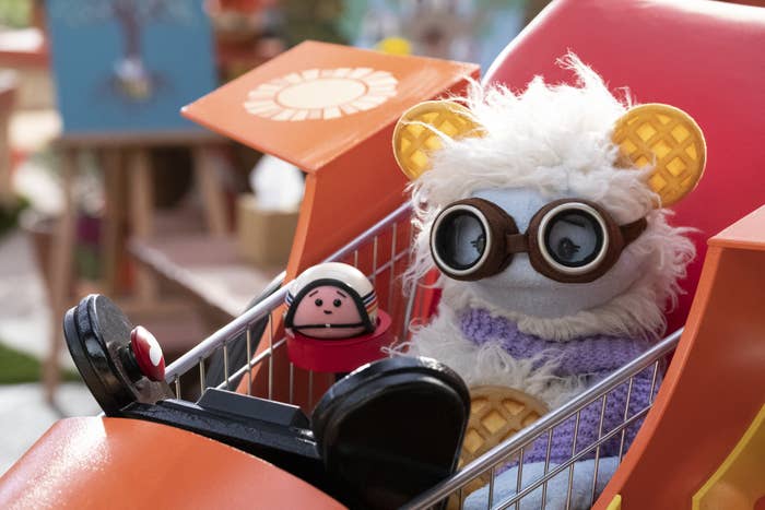 A furry puppet with frozen waffle ears, wearing protective flight goggles, is seated next to a round mochi puppet, wearing a helmet inside a flying go-kart
