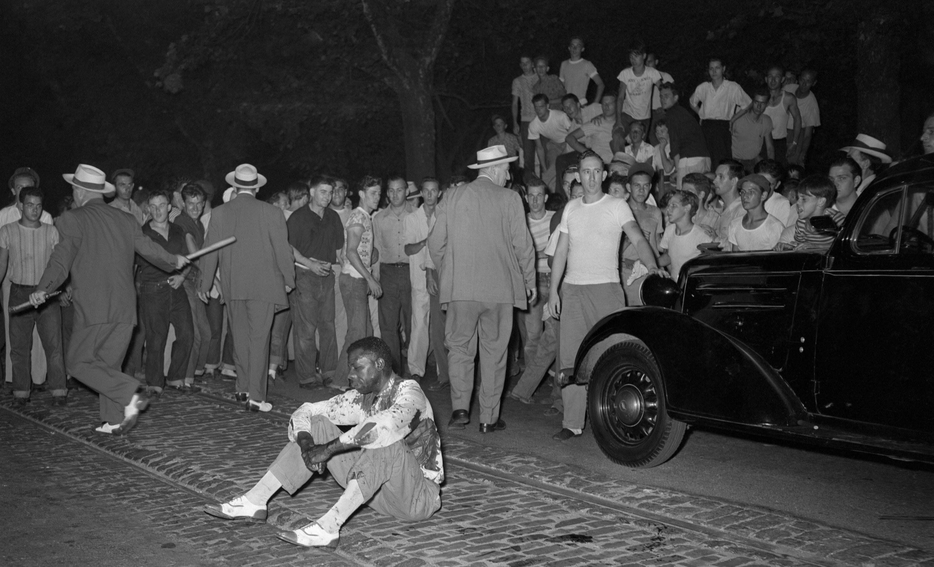 A beaten Black man sits in the street while police check members of the mob who attacked and stoned him during a race riot, which started when Black people were permitted to use public swimming pools along with whites for the first time