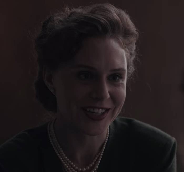 A woman is sitting at a desk, wearing pearls and smiling in a friendly manner