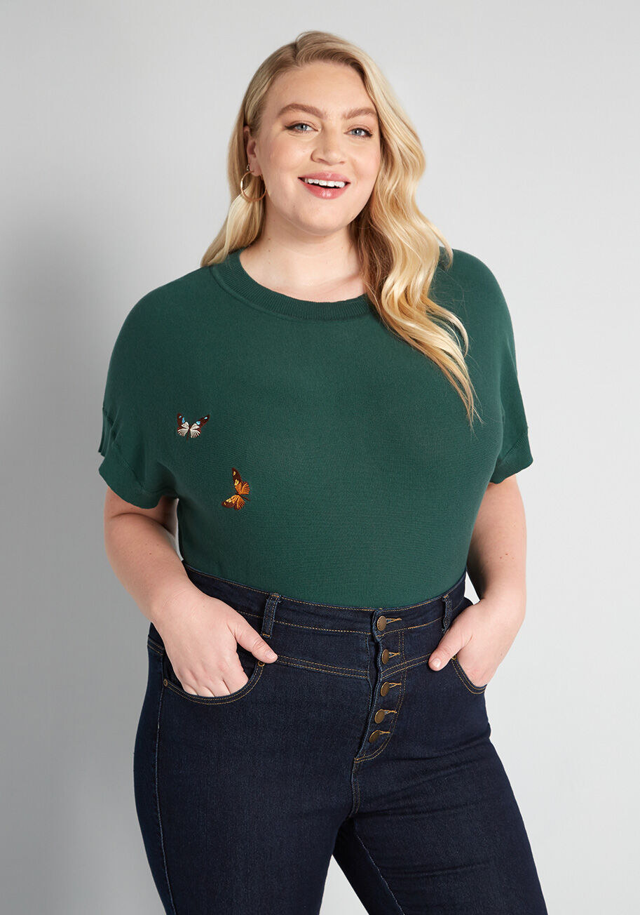 Plus size model wearing the shirt tucked into jeans. It has short sleeves and two butterflies embroidered on one side. 