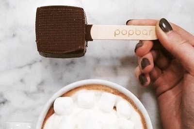 The square of chocolate on a stick held next to a mug of hot chocolate