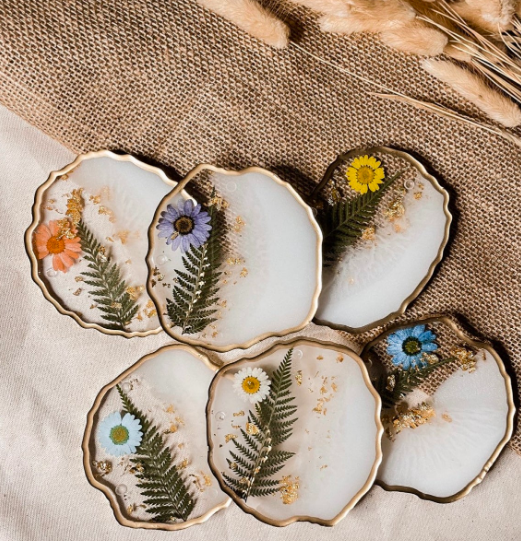 Several clear coasters with brushed white paint inside, gold leaf, fern leaves, and flower petals