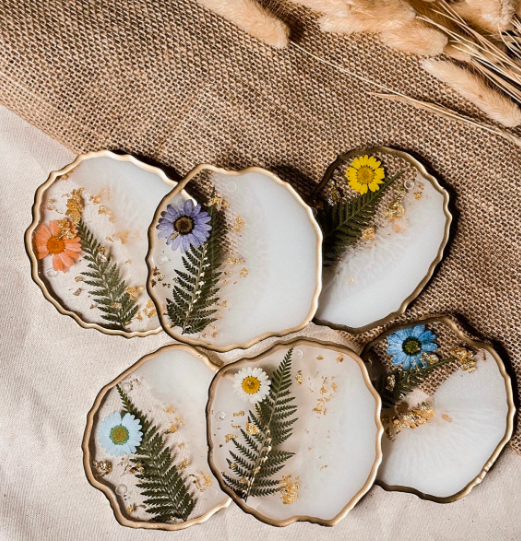 Several clear coasters with brushed white paint inside, gold leaf, fern leaves, and flower petals 