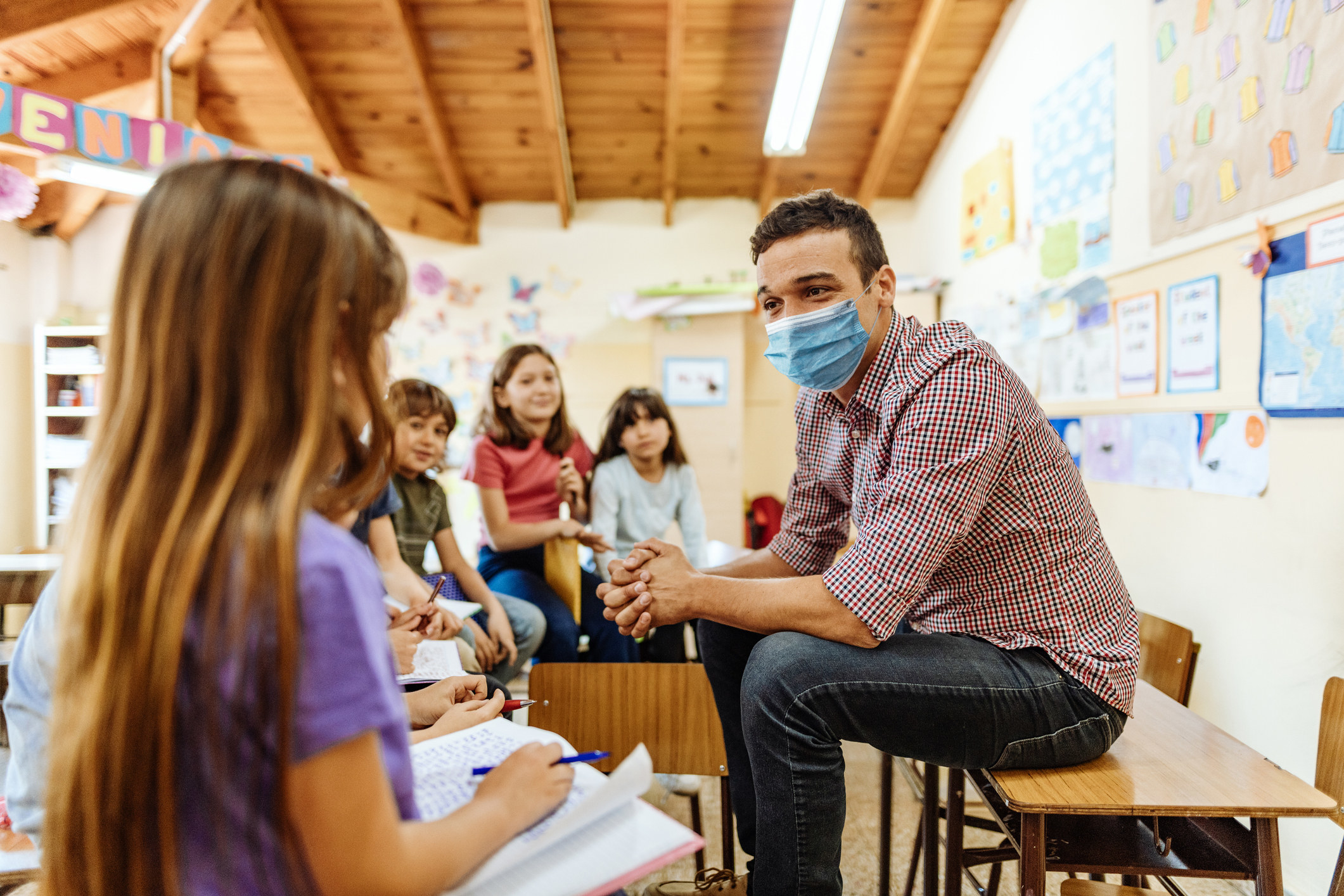 A teacher sitting on top of a desk, wearing a mask, and talking to his students