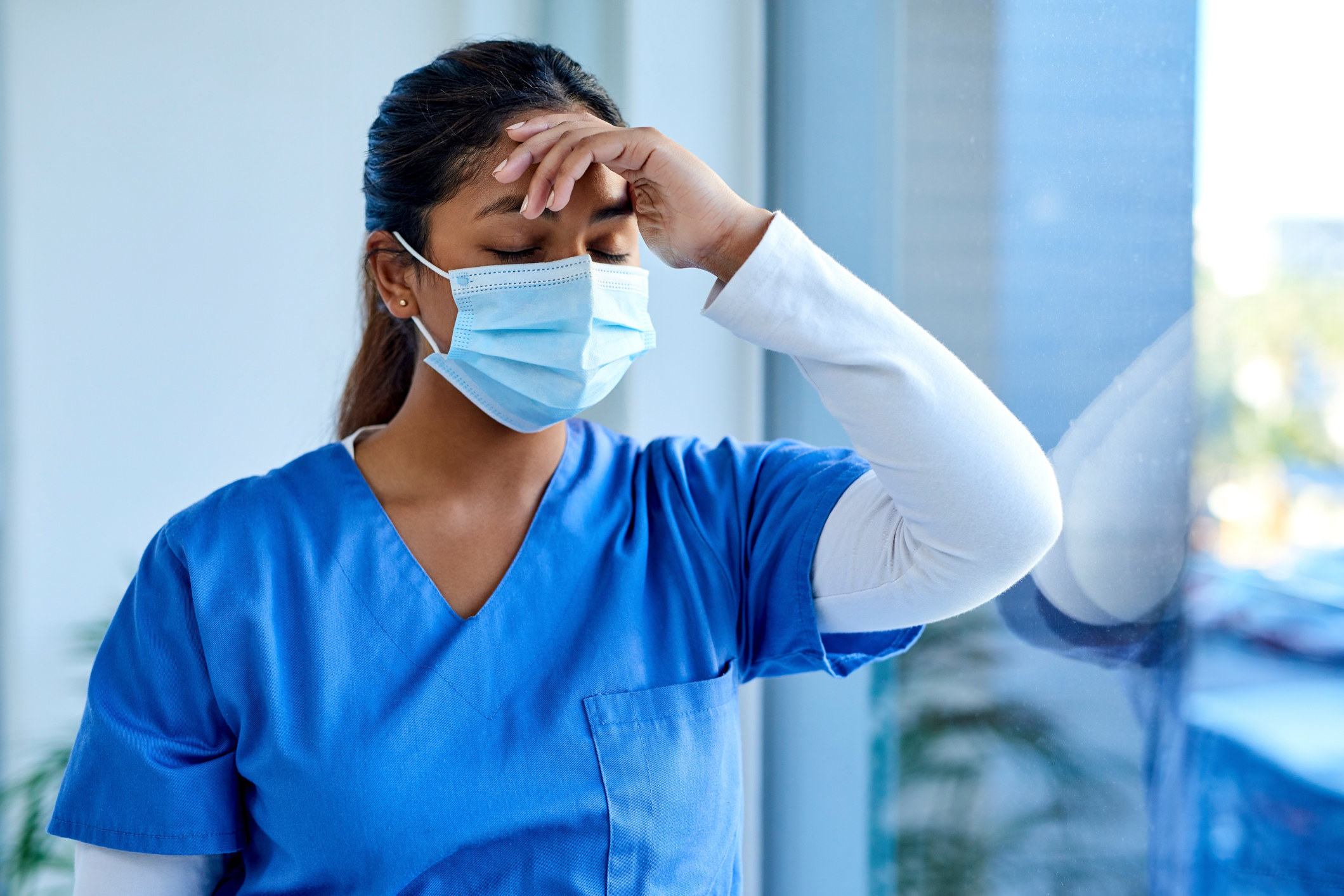 Doctor/nurse wearing mask in hospital, overworked, leaning against glass window during covid