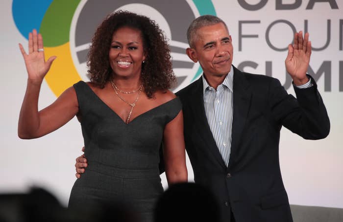 Michelle and Barack Obama stand, with Barack&#x27;s arm around Michelle&#x27;s waist, and wave at an audience