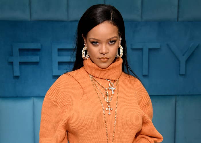 Rihanna wears an orange sweater in front of a Fenty sign