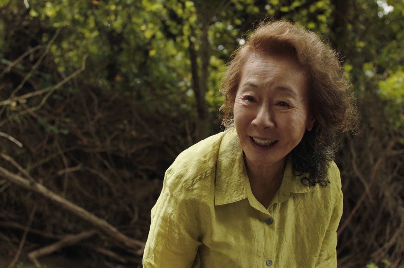 An older woman, Youn Yuh-Jung, in a yellow shirt in front of plants outdoors in a still from Minari
