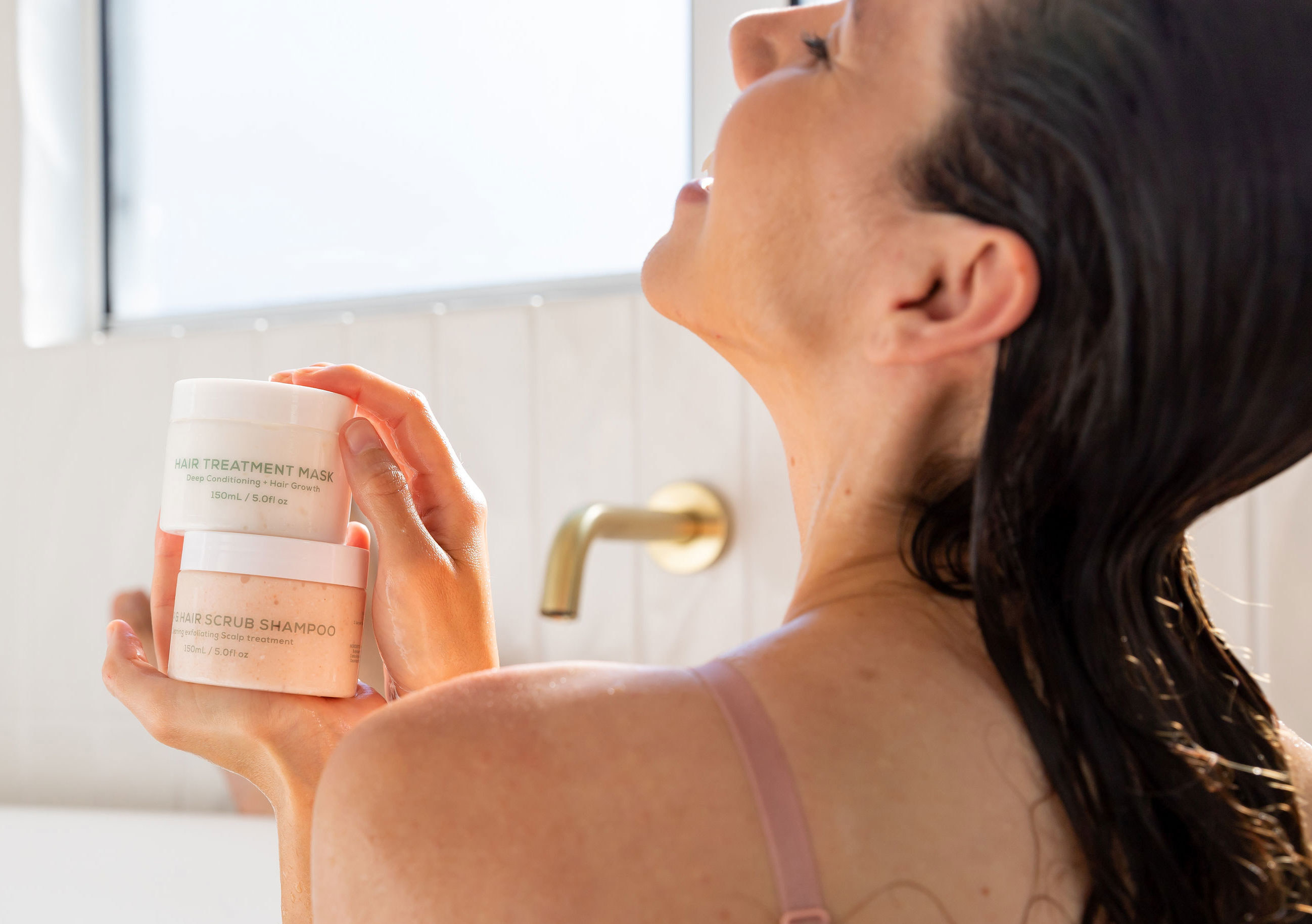 A model in a bathtub holding the containers of the scalp scrub and hair treatment mask