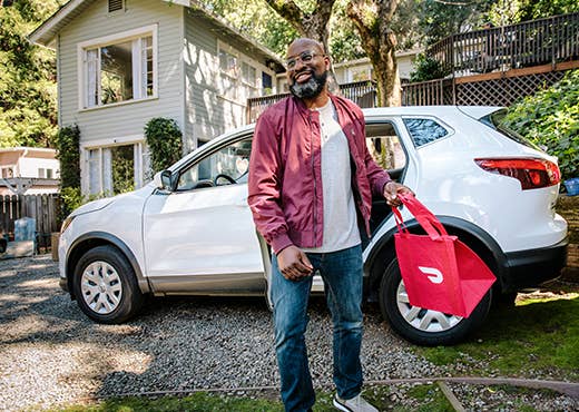 Man stands beside parked car holding a DoorDash bag. 