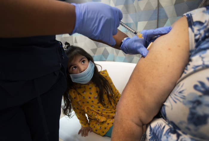 A young child looks at a needle going into the arm of an adult woman, administered by a person wearing medical gloves