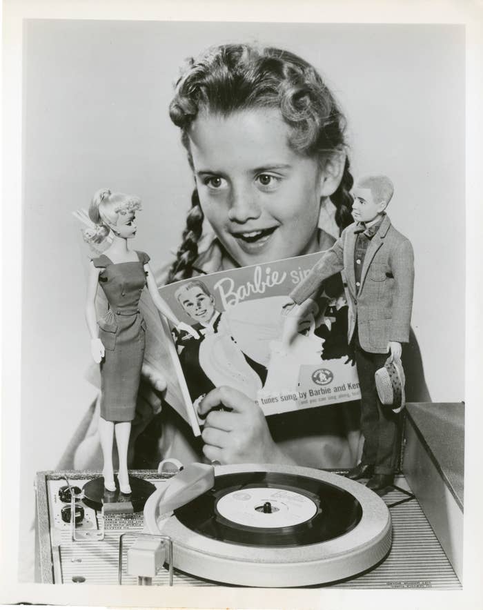 A 1960s black and white photo of a little girl in braids holding a Barbie sing along book with Barbie and Ken standing on a record player