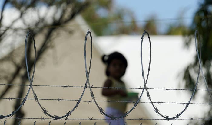 A child stands in the background, out of focus, between loops of barbed wire on a fence in the foreground