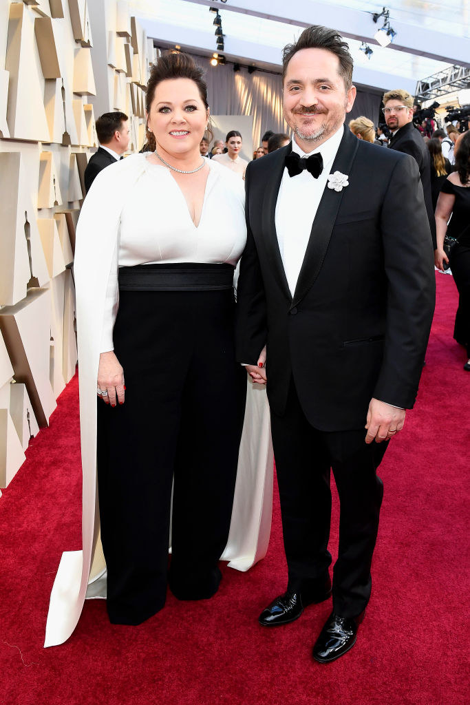 Melissa McCarthy in a black-and-white caped outfit with pearls around her neck and Ben in a tuxedo on the red carpet  at the 91st Annual Academy Awards