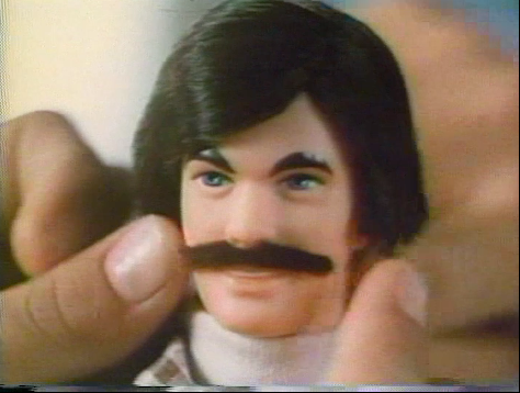 A screen shot of a hand putting a mustache on a Ken doll