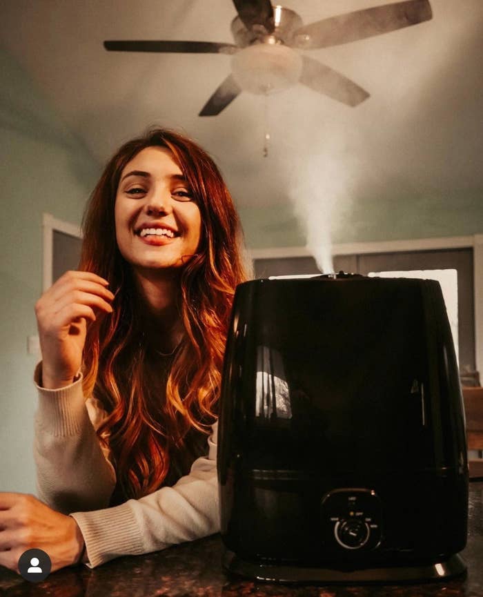 A reviewer smiles next to their black humidifier while it dispenses a white fog