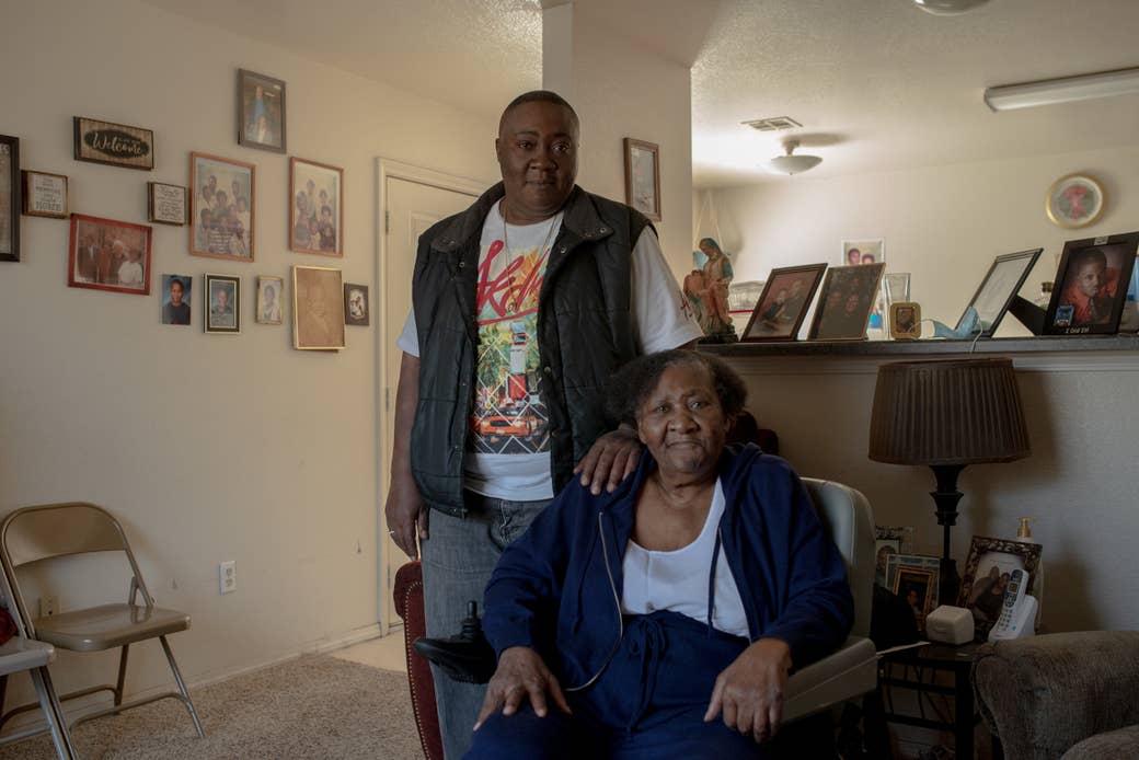 A son stands behind his mother, who is in a motorized wheelchair, as both pose in their living room, which has several family portraits on the walls and counter behind them