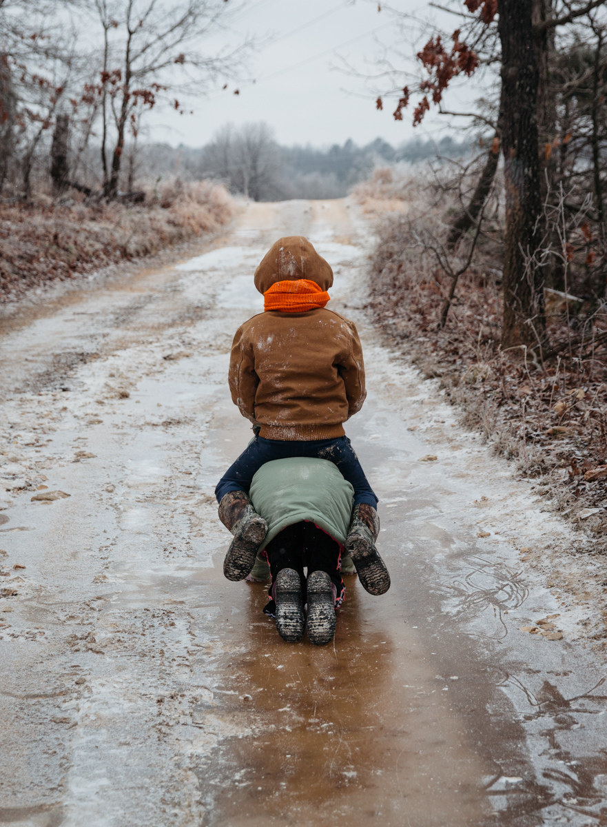 A child sits on the back of another child, who is on their knees on an icy muddy path 