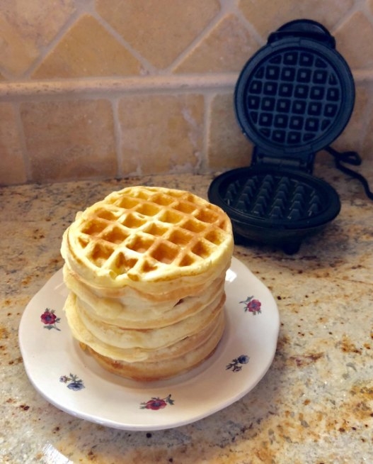 A tiny stack of waffles sit on a floral plate. A small black waffle maker sits open just behind the plate.