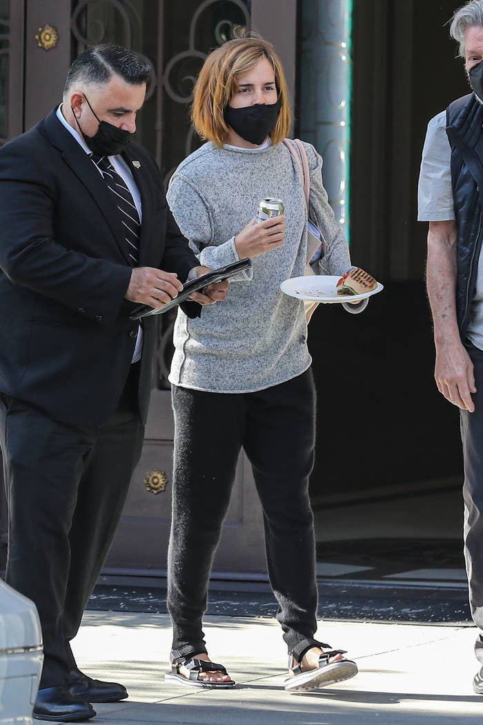 Emma with a chin-length bob, wearing comfortable-looking pants, sweater, sandals, and a face mask and holding a beverage can and a plate with a sandwich