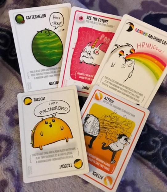 Five Exploding Kittens cards laid out including a TacoCat card, a CatterMelon card, and a Rainbow-Ralphing Cat. 