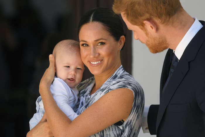 Meghan holds a baby and smiles at the camera as Harry stands next to her