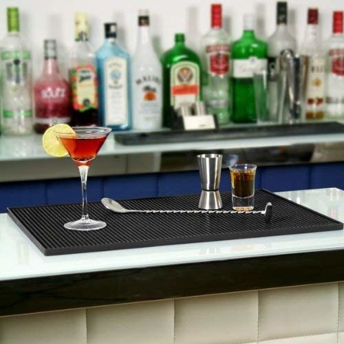 A black PVC bar mat pictured with a glass of cocktail and other bar accessories on top of it
