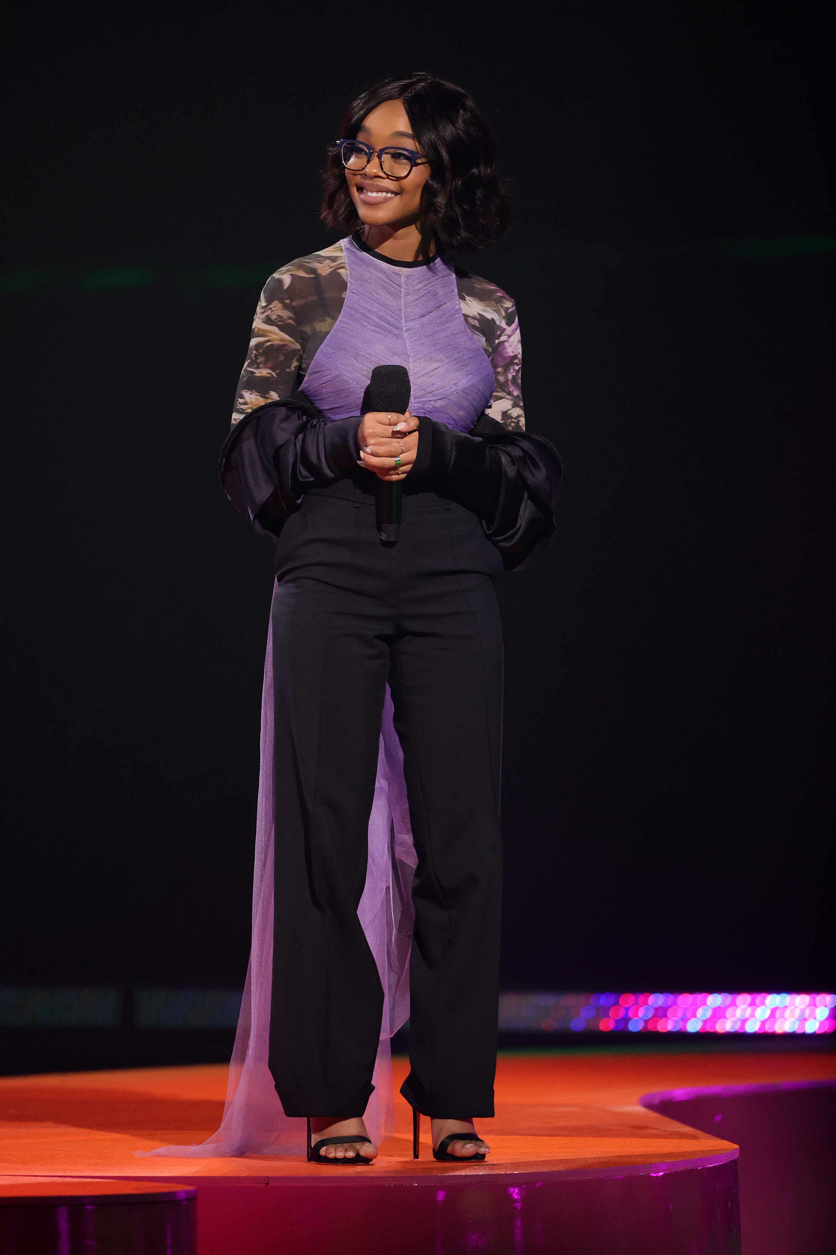 Bespectacled Marsai smiling and wearing high heels, black pants, and a top with flowy sleeves and a purple train