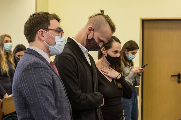 Standing among a row of people wearing face masks in a courtroom, Lang looks to the ground and Ospiovich puts a hand to her neck