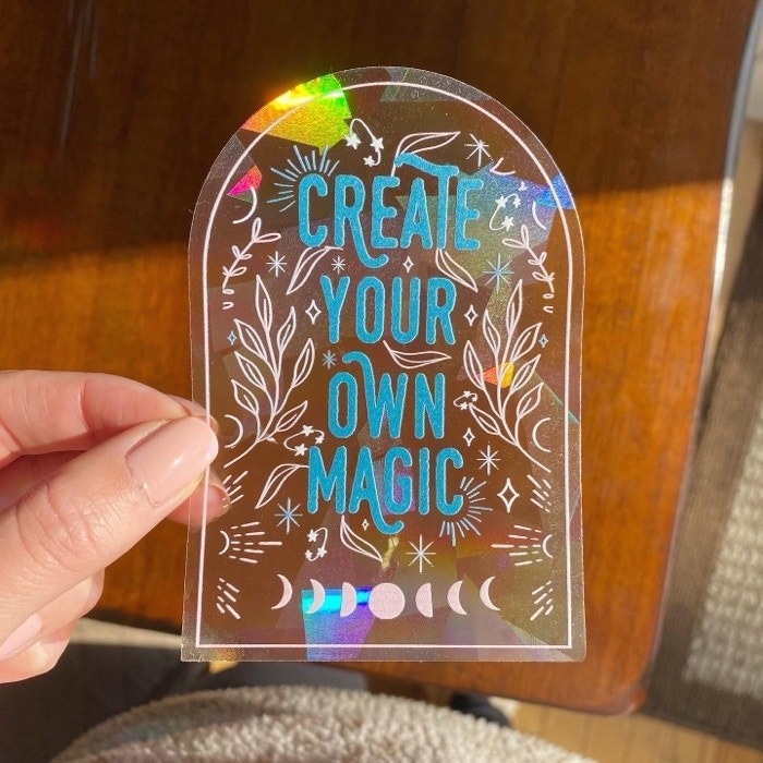 A hand holding a window decal suncatcher that says &quot;Create your own magic&quot; 