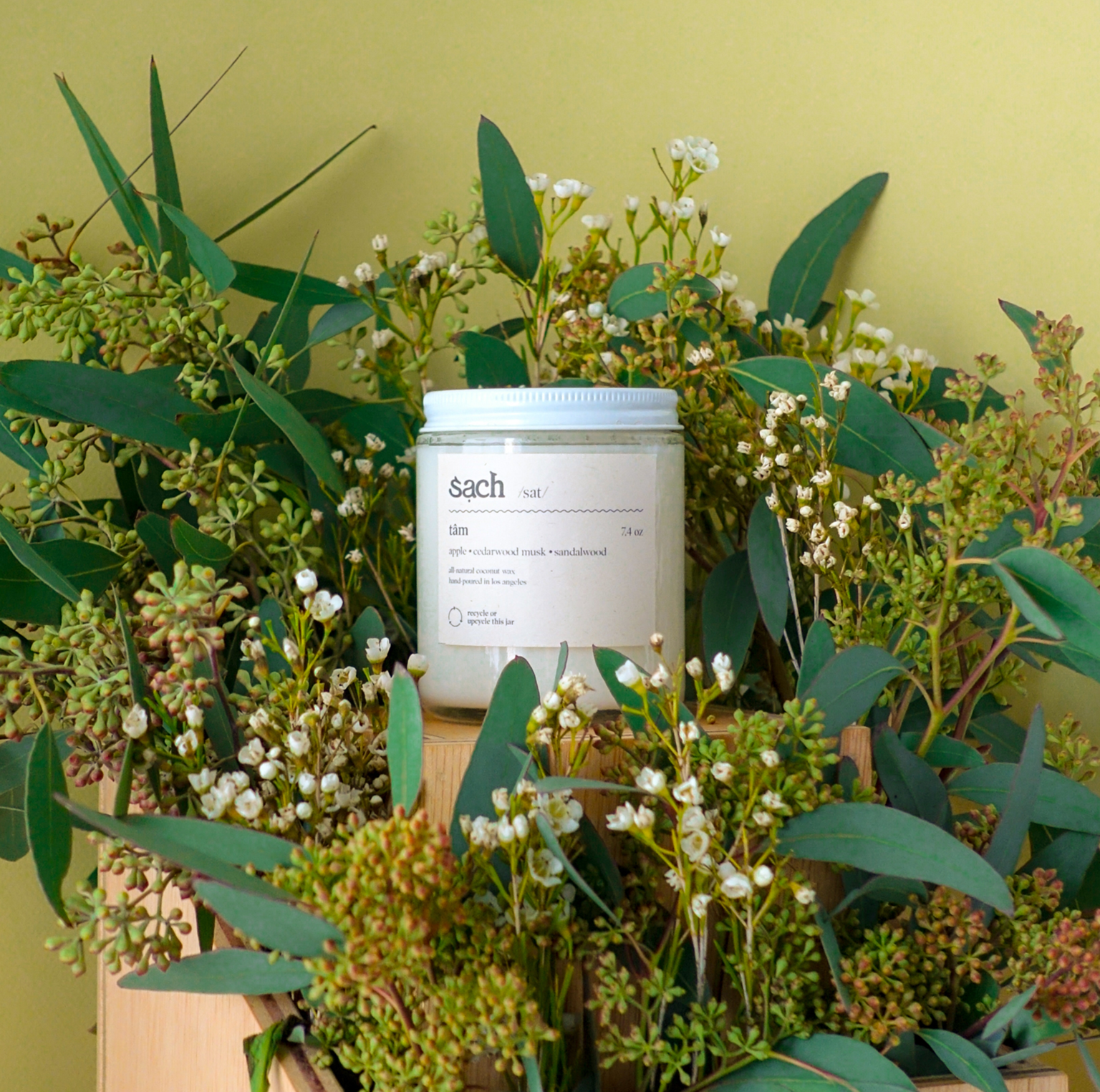 A white candle in a screw-top jar surrounded by green flowers in brow paper bags 