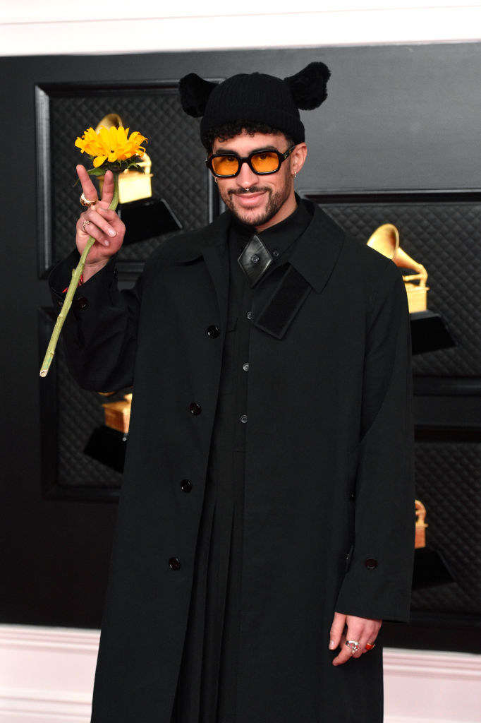 Bad Bunny wearing an all-black outfit, sunglasses, a beanie and holding a sunflower