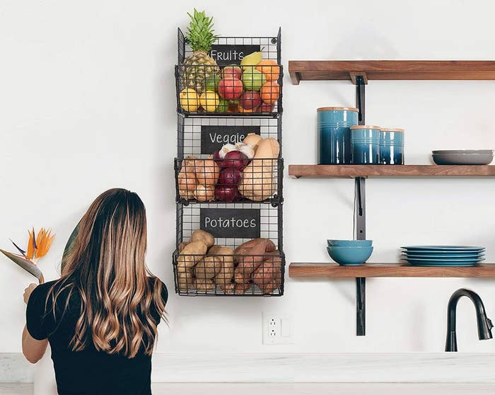 A person standing in front of the three-tiered basket shelf mounted to a wall full of produce 