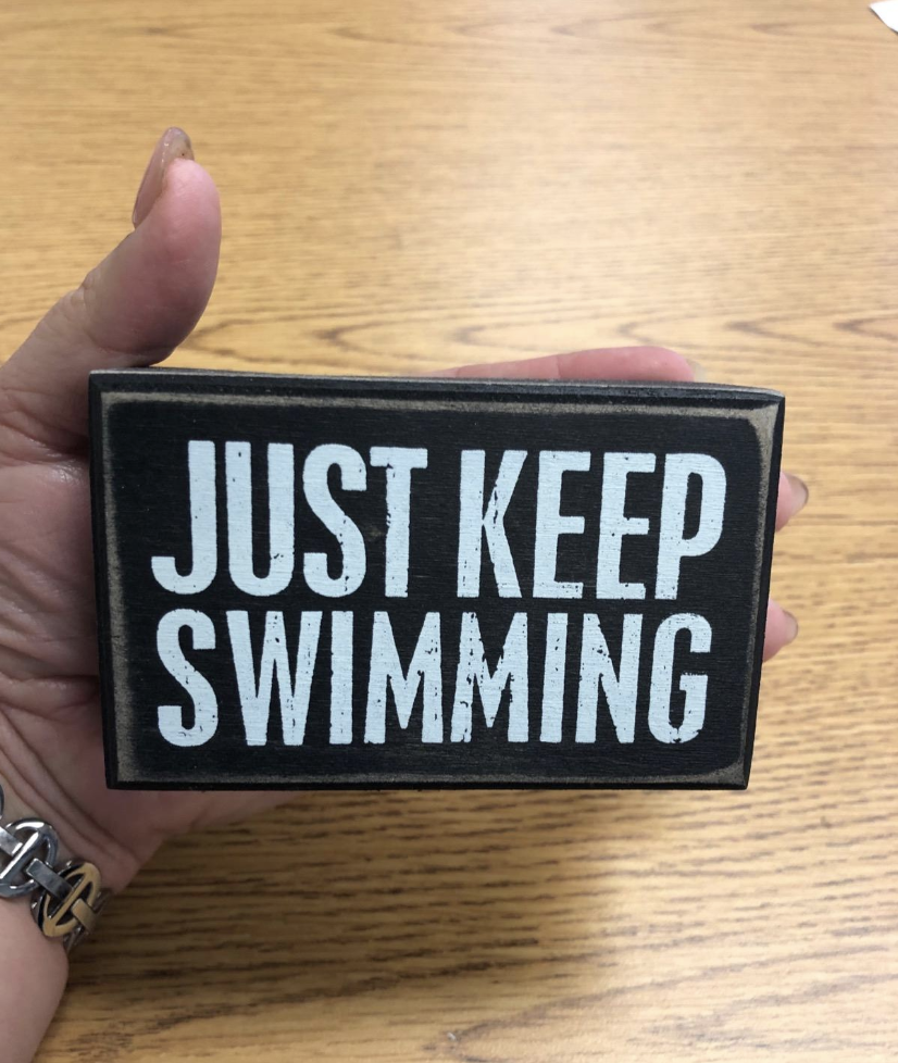 Reviewer holding hand-sized brown box sign that says &quot;Just Keep Swimming&quot; in white block letters 