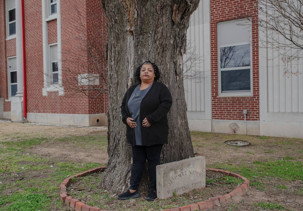 A woman stands in front of a tree, marked with a cement sign that says &quot;Seminole Nation Whipping Tree&quot;