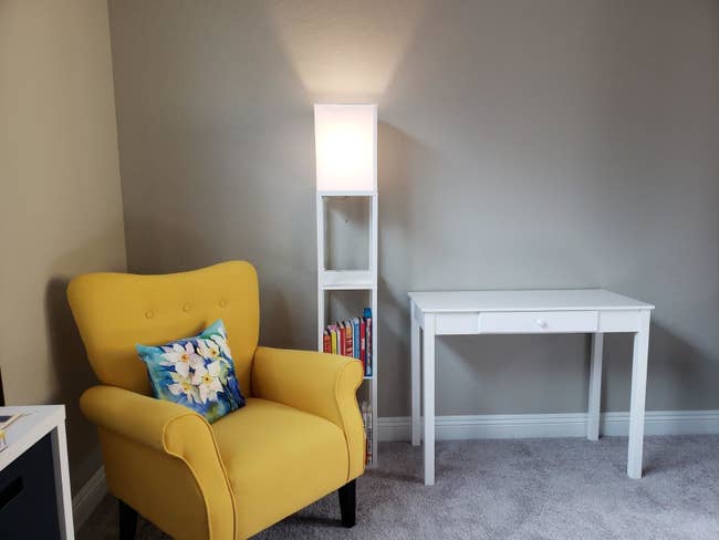 reviewer image of the white brightech maxwell charger shelf floor lamp turned on in a reading nook next to a yellow armchair