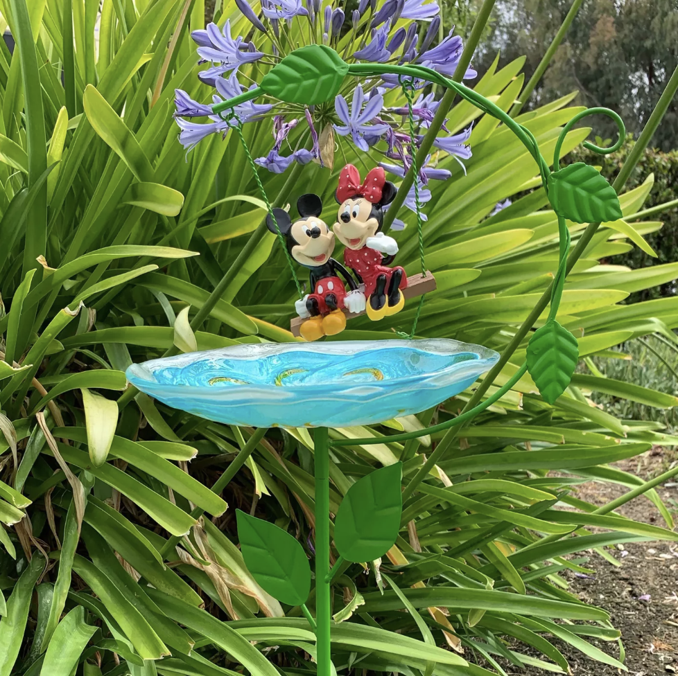 A ground-fixed bird bath with a blue dish and Mickey and Minnie figures on a swing hovering over it 