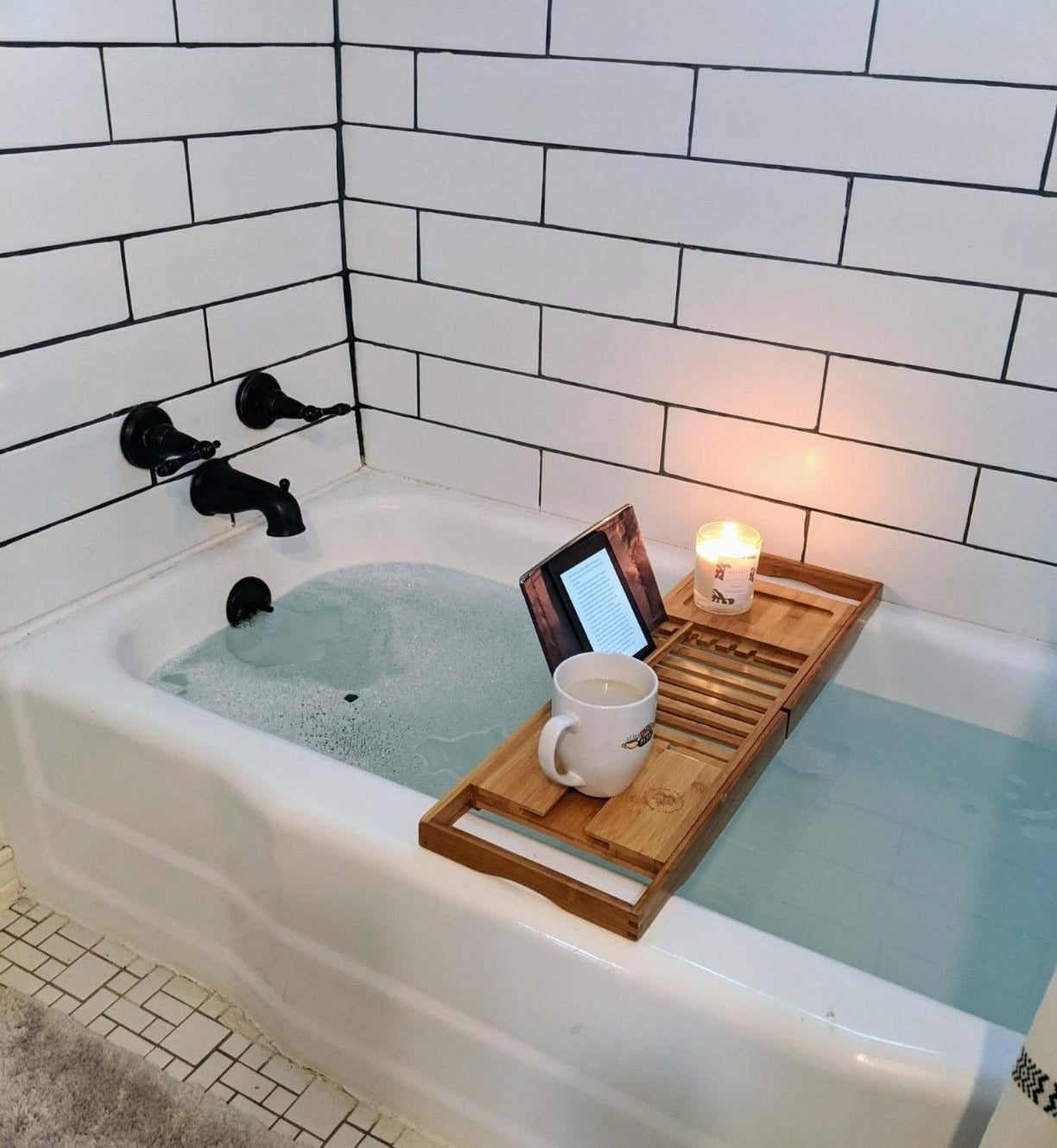 wooden caddy stretched over a tub holding a propped-up kindle, a candle, and a mug