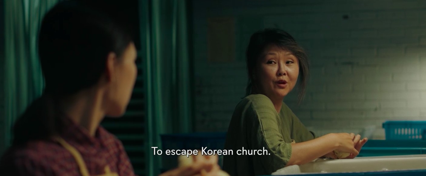 Monica sitting down at her chicken-sexing job, conversing with her coworker. Text subtitles on image read, &quot;To escape Korean church.&quot;