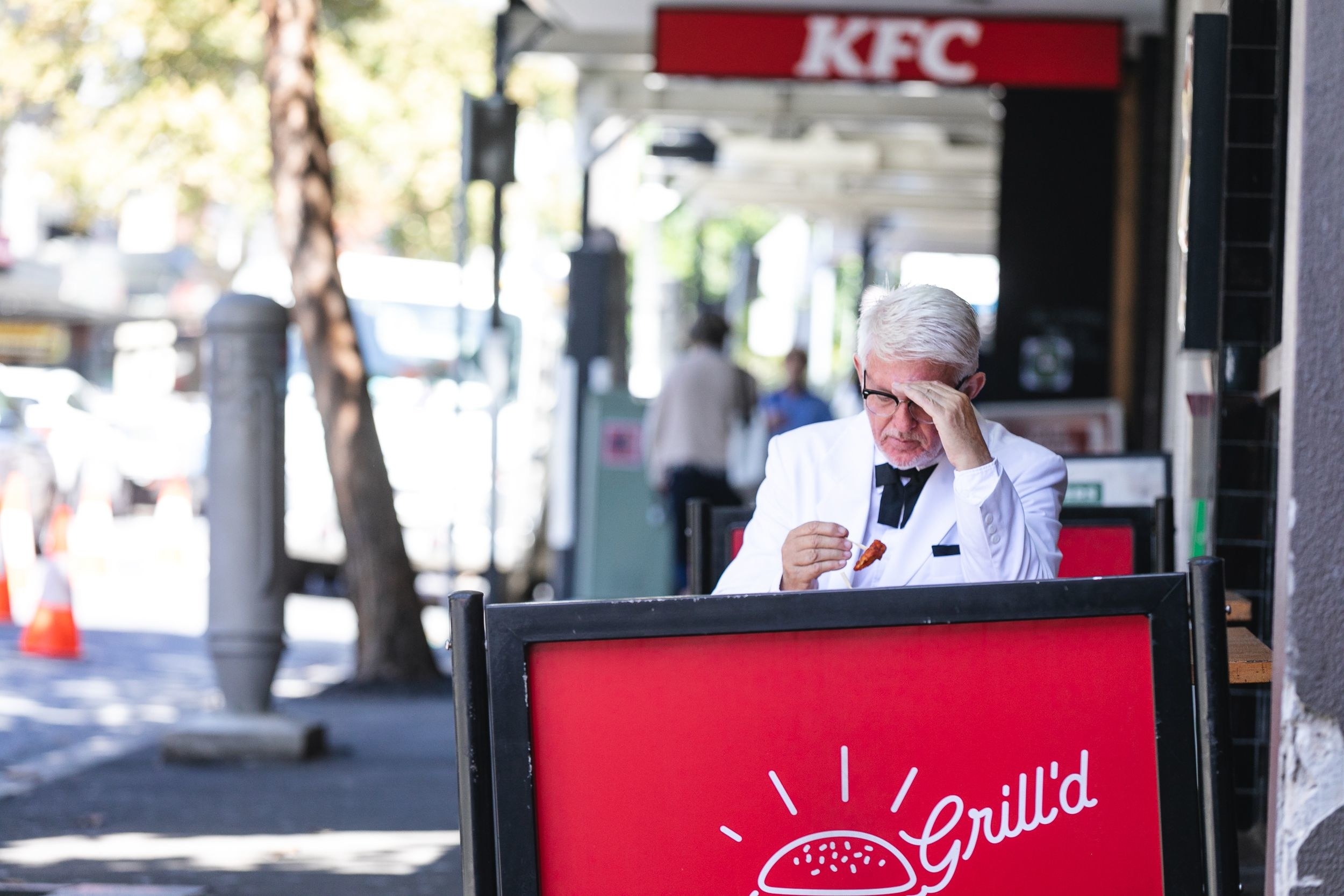 Impersonator eating Grill&#x27;d outside with a KFC store sign perfectly clear in the background
