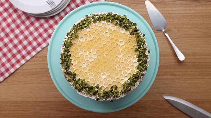 An overhead shot of a cake on a blue cake-stand with red and white checkered cloth on a wood table with utensils around it.