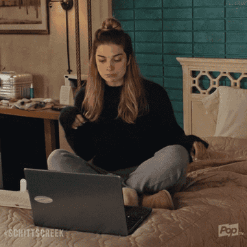 Alexis Rose patting a spot next to her on a bed with a laptop in front of her 