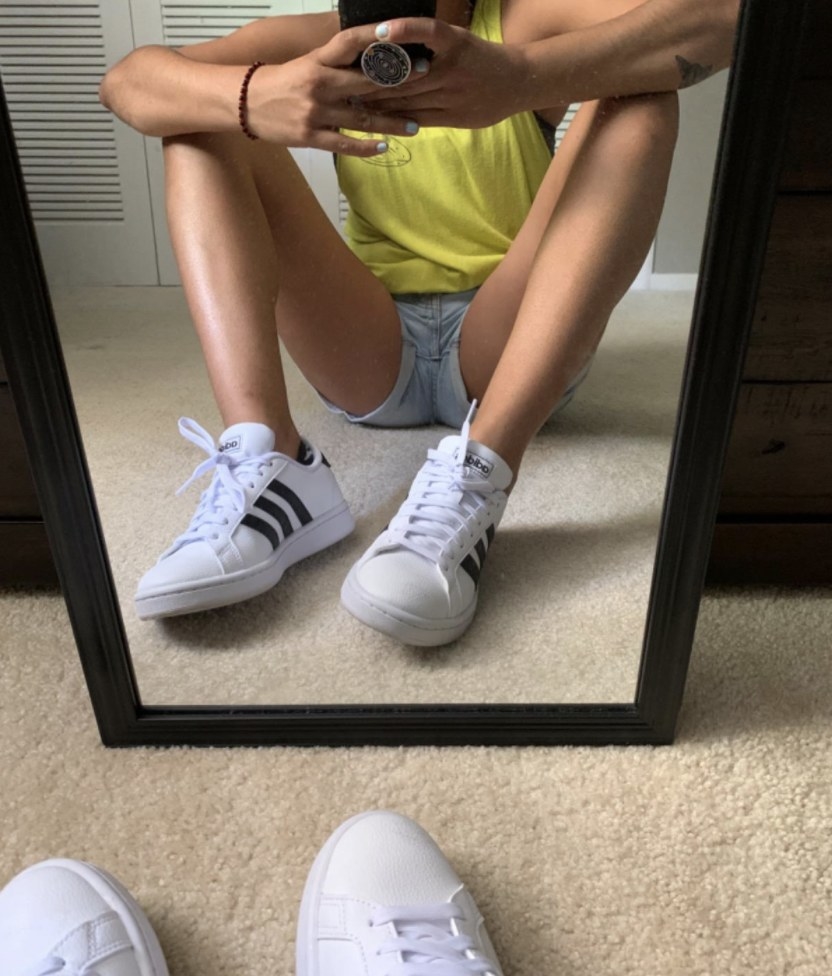A person is sitting in front of a mirror wearing white shoes with three black stripes on the side