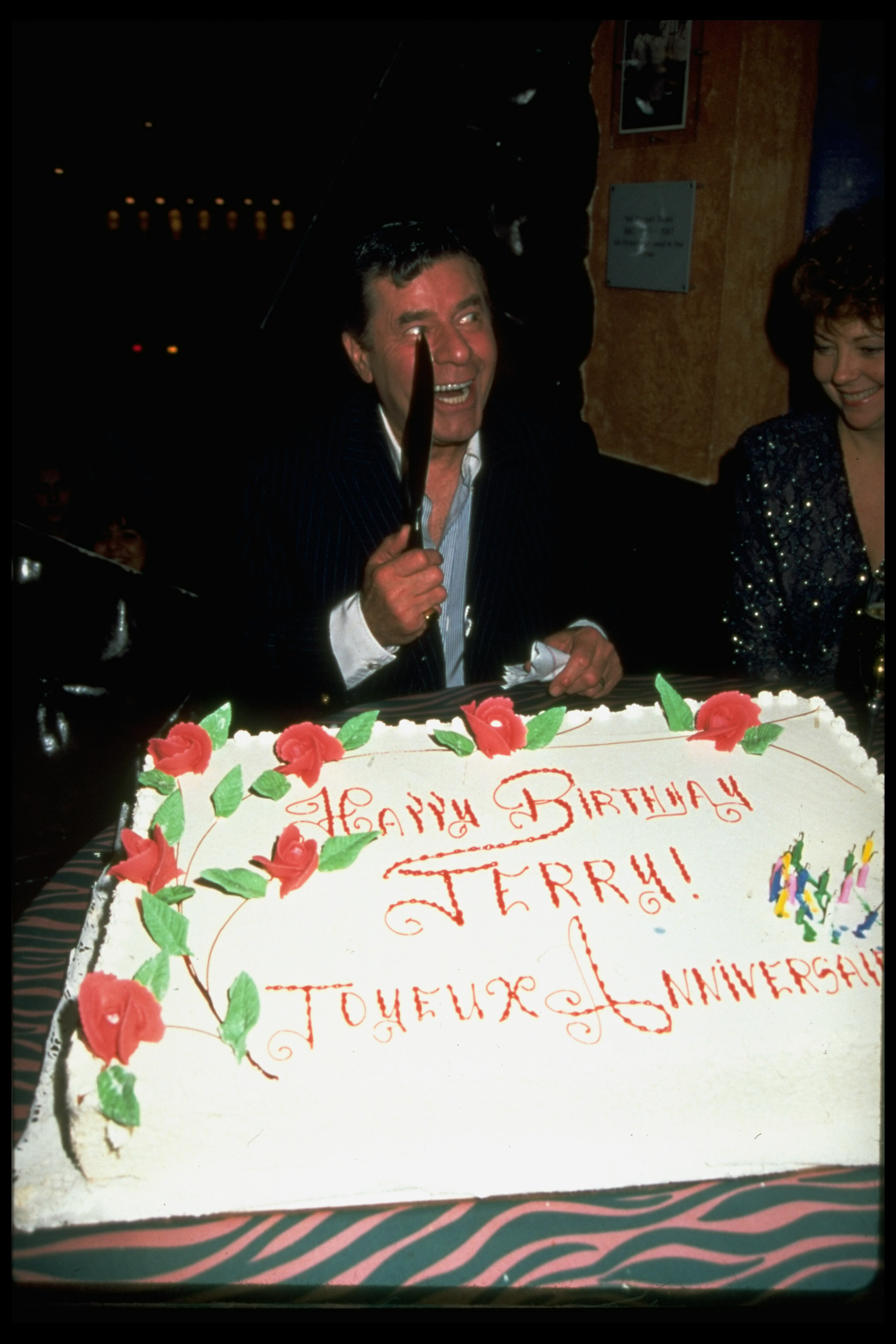 Jerry Lewis holds a knife jokingly in the air over a big cake that reads Happy birthday Jerry in french and english 