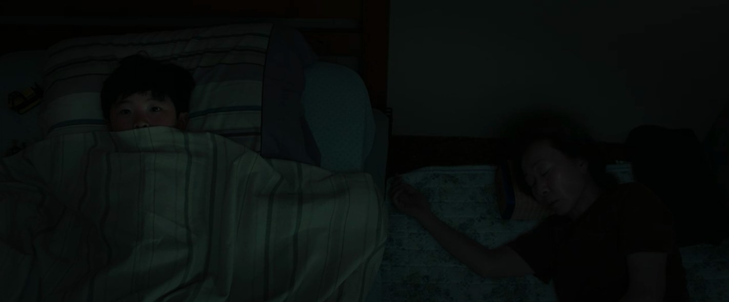 David lying on his bed in the dark with his blanket over his face, while his grandma sleeps on the floor directly to his left.